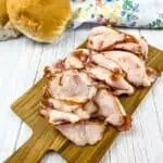 Sliced pork lunch meat on a cutting board with buns in the background.