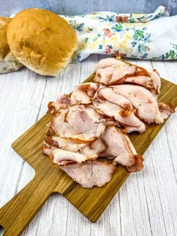 Sliced pork lunch meat on a cutting board with buns in the background.