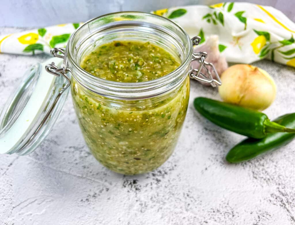 Tomatillo sauce in a glass jar with veggies in the background.