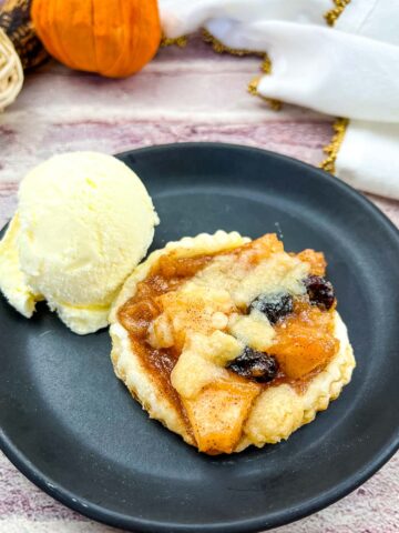 An apple-cranberry puff pastry tart on a black plate with a scoop of ice cream.