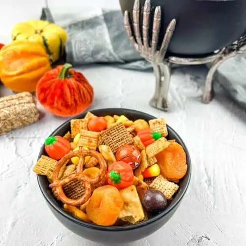 Spicy chex mix in two black bowls with pumpkins in the background.