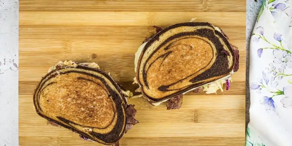 Two Reuben sandwiches made with marbled rye bread sit on a wooden cutting board next to a floral-patterned cloth, ready to be enjoyed alongside a classic Rachel sandwich.