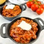 A closeup shot of Smoked Brisket Chili in 2 black bowls with bread and tomatoes in the background.