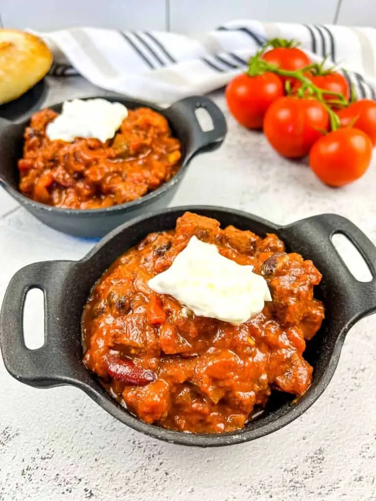 A closeup shot of Smoked Brisket Chili in 2 black bowls with bread and tomatoes in the background.