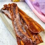 Smoked candied bacon on a plate with maple syrup in the background.