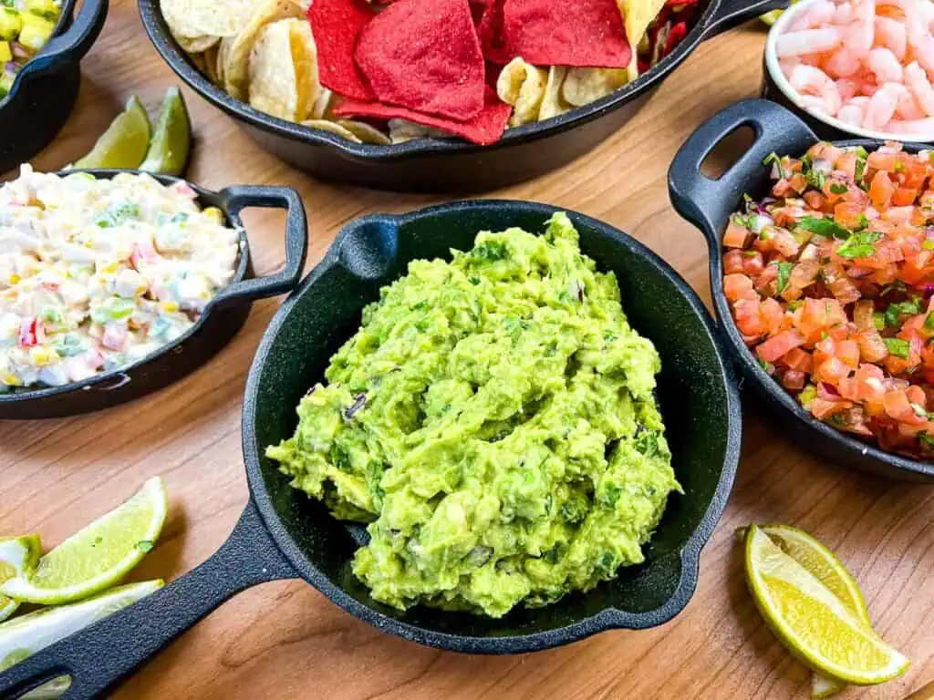 A bowlful of guacamole on a guacamole board with salsas and chips in the background.