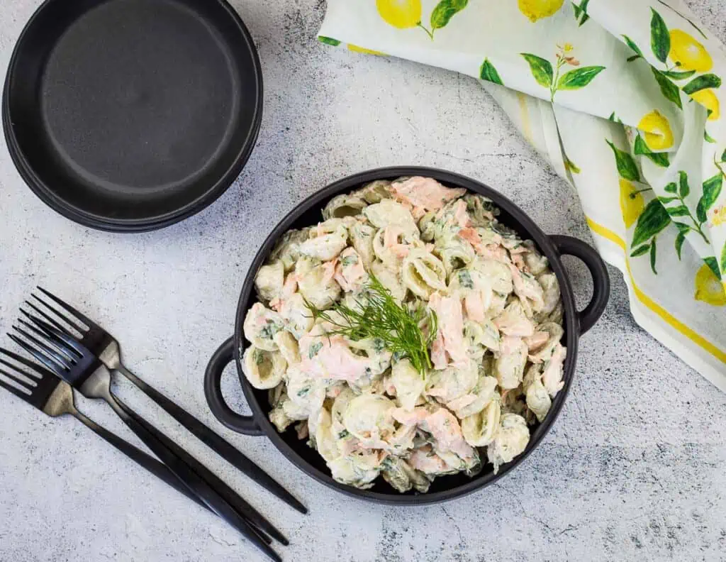 Salmon pasta salad in a black bowl with forks and plates nearby.