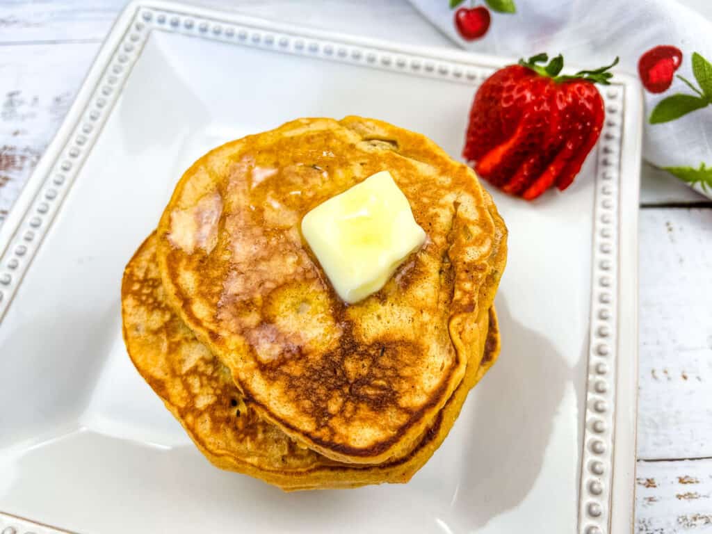 A stack of Sweet Potato Pancakes with pecans on a plate with a strawberry in the background.