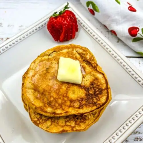 A stack of Sweet Potato Pancakes with pecans on a plate with a strawberry in the background.