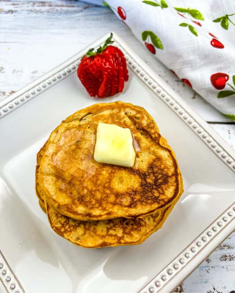 A stack of Sweet Potato Pancakes with pecans on a plate with a strawberry in the background.