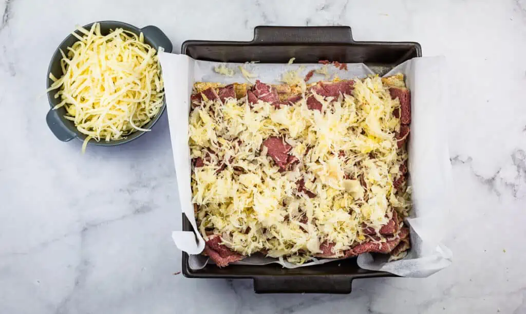 A square baking pan lined with parchment paper holds uncooked layered Reuben sliders topped with shredded cheese, next to a bowl of extra cheese on a marble surface.