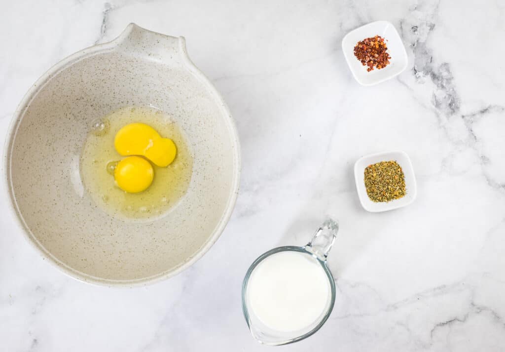 Cracked eggs in a bowl next to milk and spices.