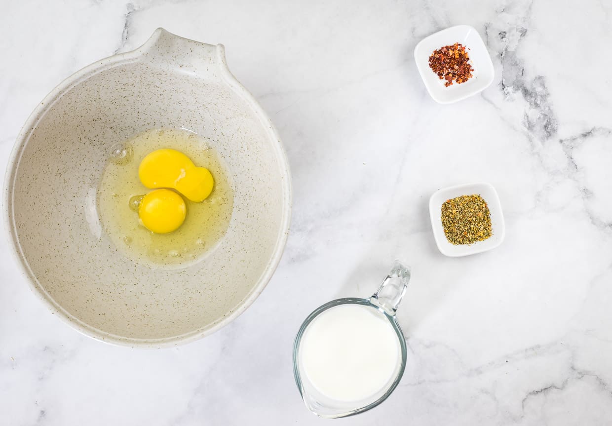 Cracked eggs in a bowl next to milk and spices.