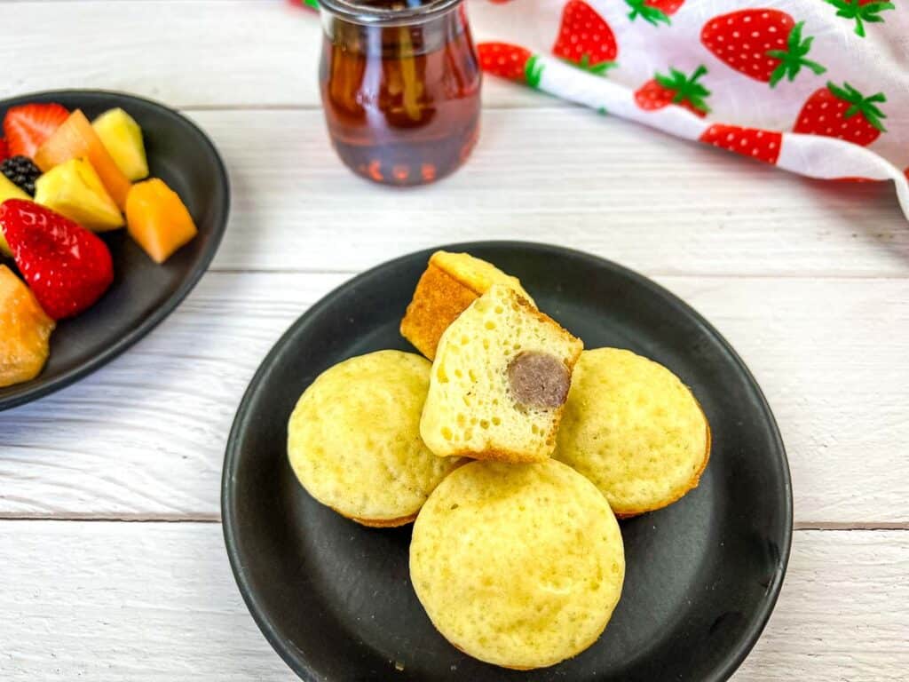 Pancake bites with sausage on a black plate with fruit and syrup in background.