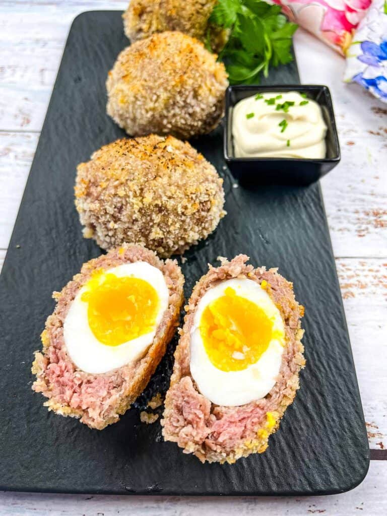 Cut open Scotch Eggs and whole Scotch Eggs on a black platter.