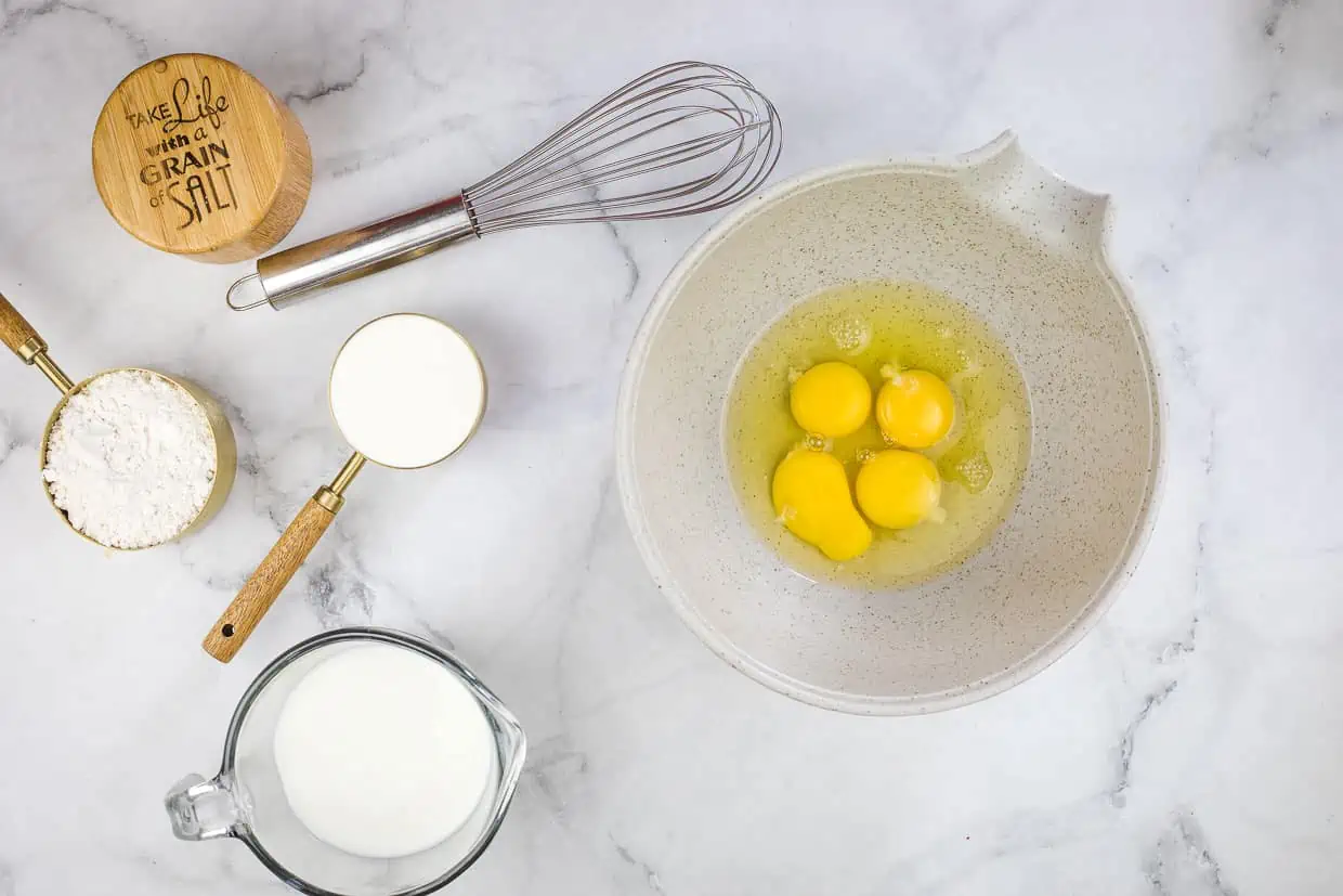 Eggs in a bowl next to a whisk and other ingredients.