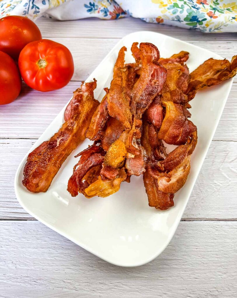 Crispy Blackstone Bacon on a white plate with tomatoes in the background.