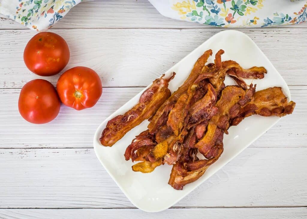 Bacon strips on a white plate next to tomatoes.