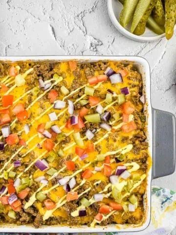 Cheeseburger Casserole in a baking dish with pickles and cheese nearby.