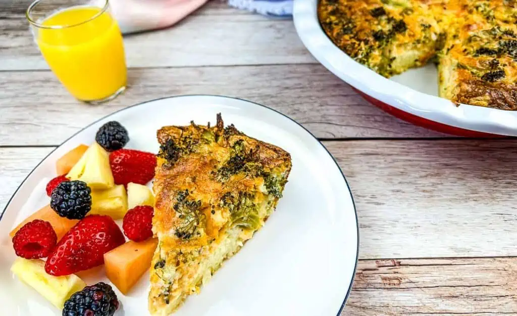 A slice of Broccoli 3-Cheese Impossible Quiche with fruit and the rest in the background.