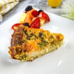 A slice of broccoli & 3-cheese quiche on a plate with fruits.
