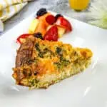 A slice of broccoli & 3-cheese quiche on a plate with fruit.