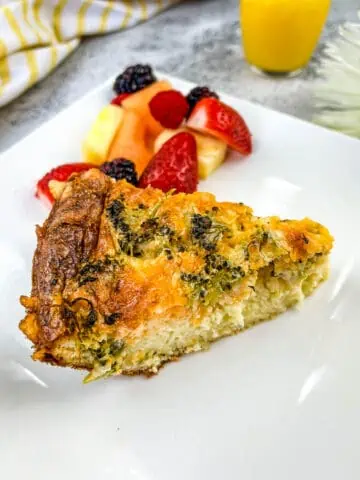 A slice of broccoli & 3-cheese quiche on a plate with fruit.