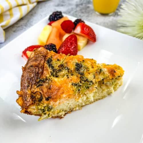 A slice of broccoli & 3-cheese quiche on a plate with fruits.