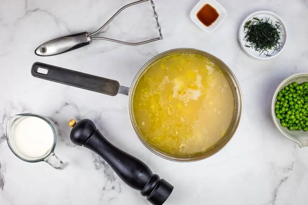 Soup with some partially mashed potatoes in it with remaining ingredients nearby.