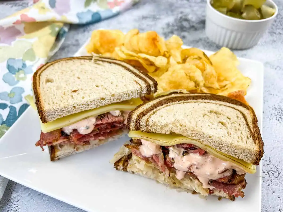 A Pastrami Reuben sandwich on a plate with chips and pickles.