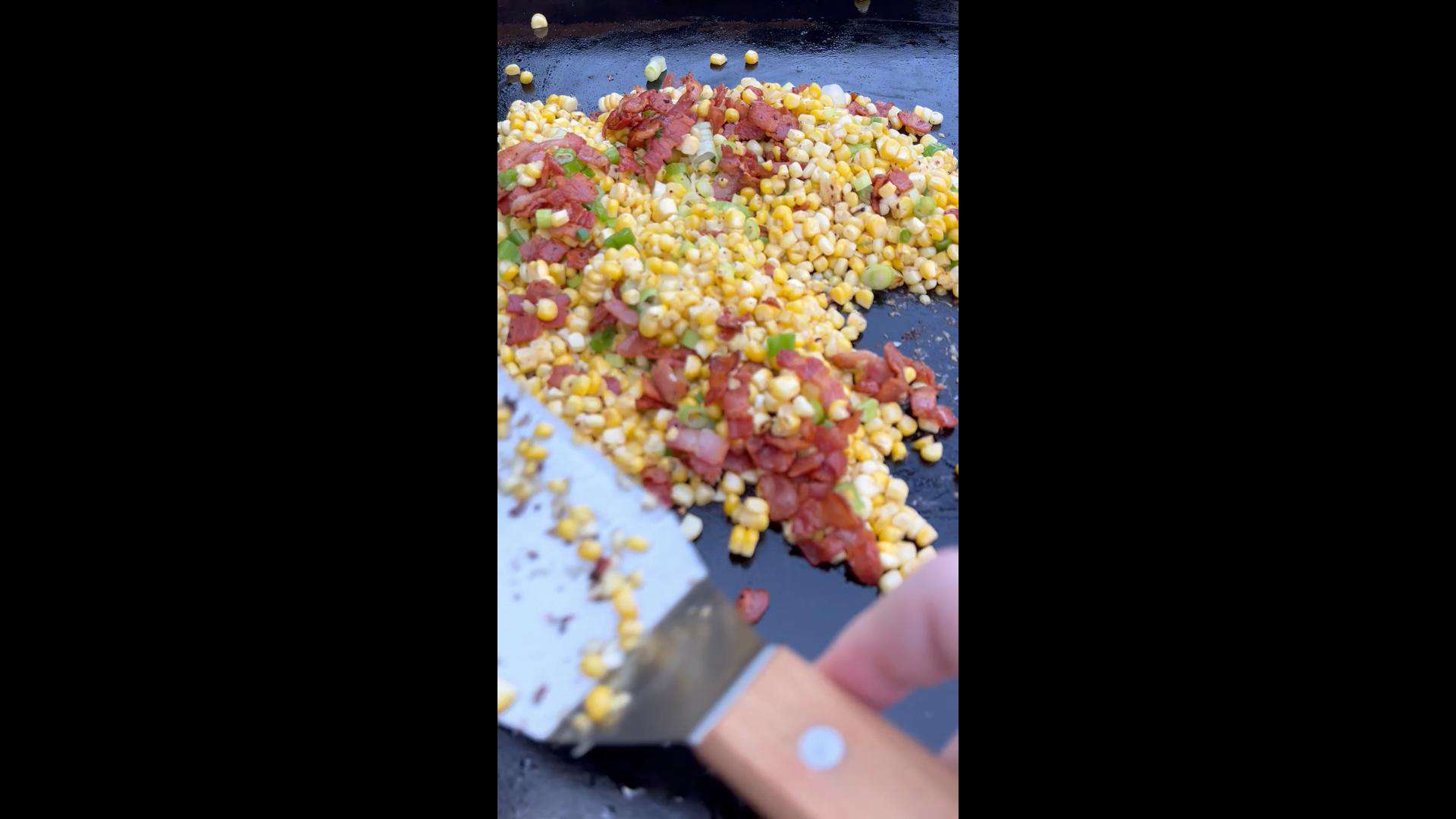 Working the cooked bacon into the corn on the griddle.
