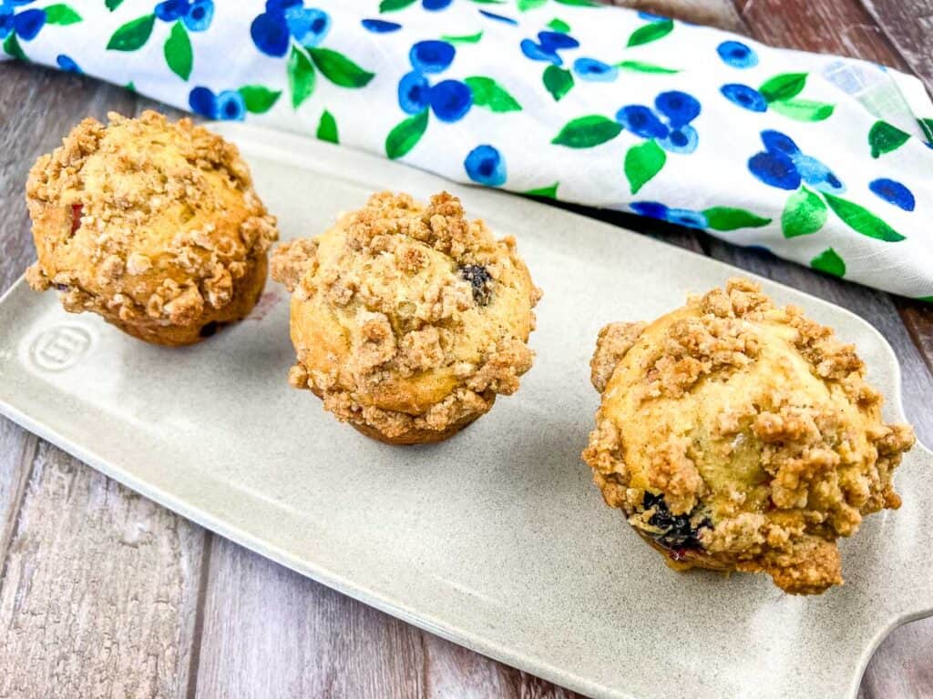 A horizontal shot of 3 blueberry-rhubarb muffins on a white platter.