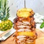 Smoked Chicken Al Pastor on a vertical skewer with pineapple in the background.