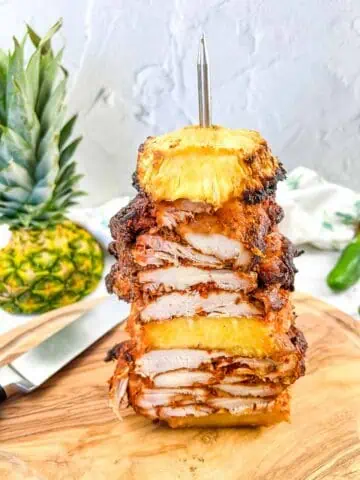 Smoked Chicken Al Pastor on a vertical skewer with pineapple in the background.