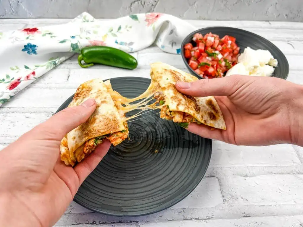 A horizontal shot of two hands pulling the quesadilla apart with gooey cheese in the middle.