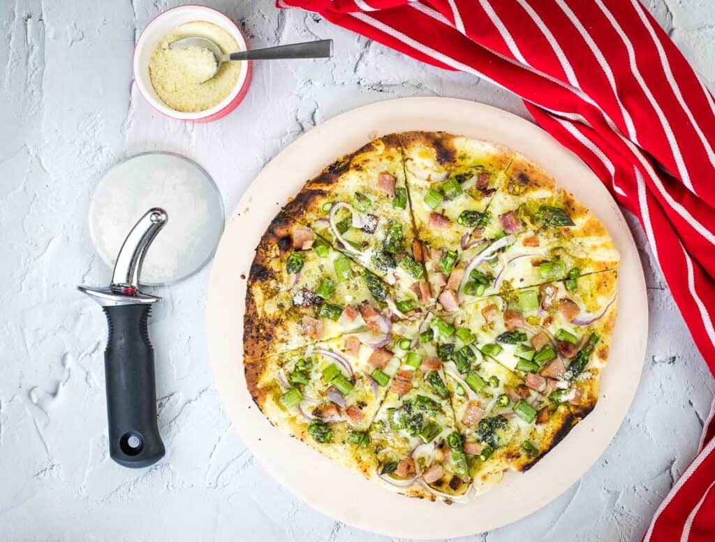 A horizontal shot of Ham and Asparagus Pizza with a pizza cutter and cheese nearby.