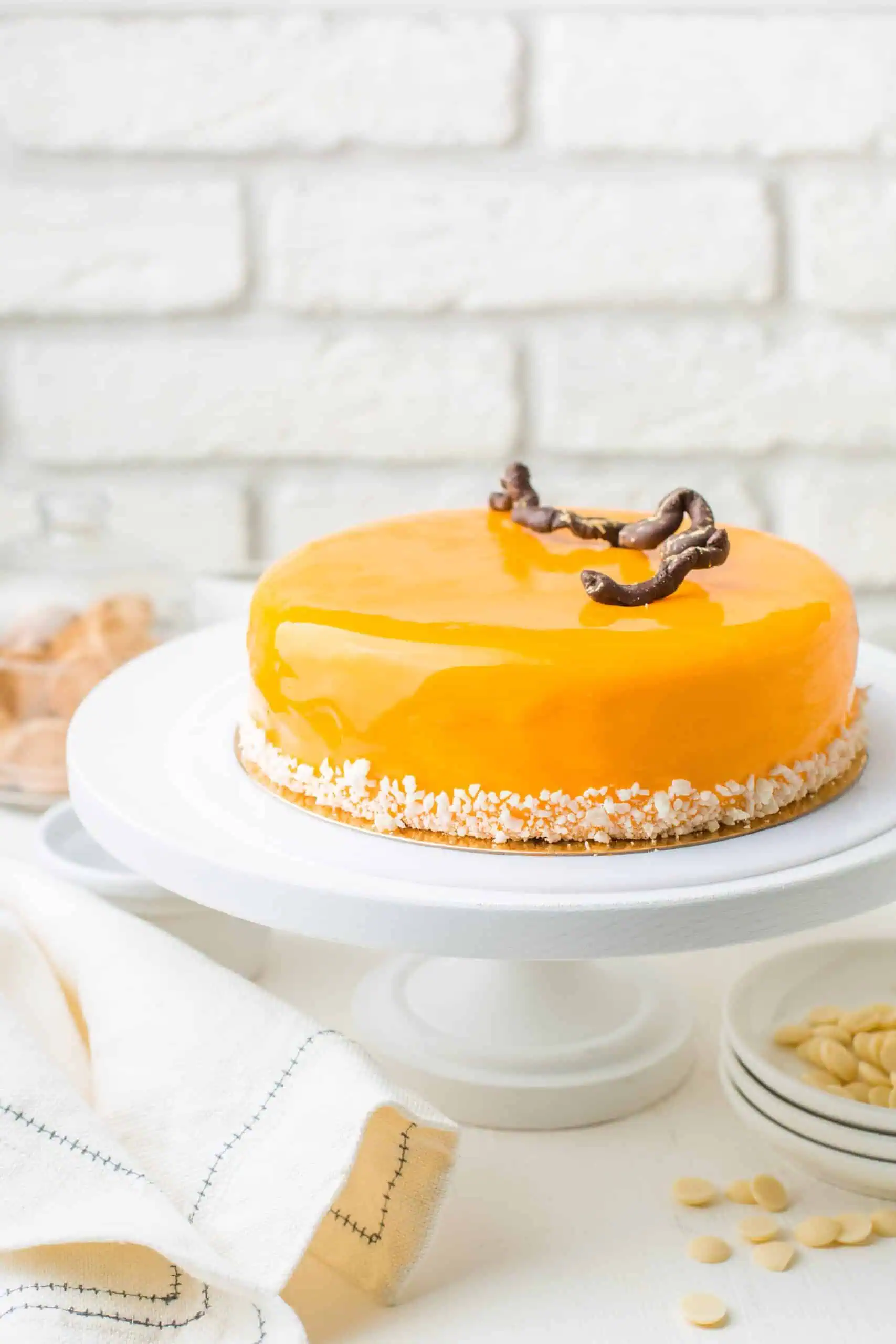 A round cake with a shiny yellow glaze sits on a white cake stand, decorated with chocolate garnish and white sprinkles around the base.