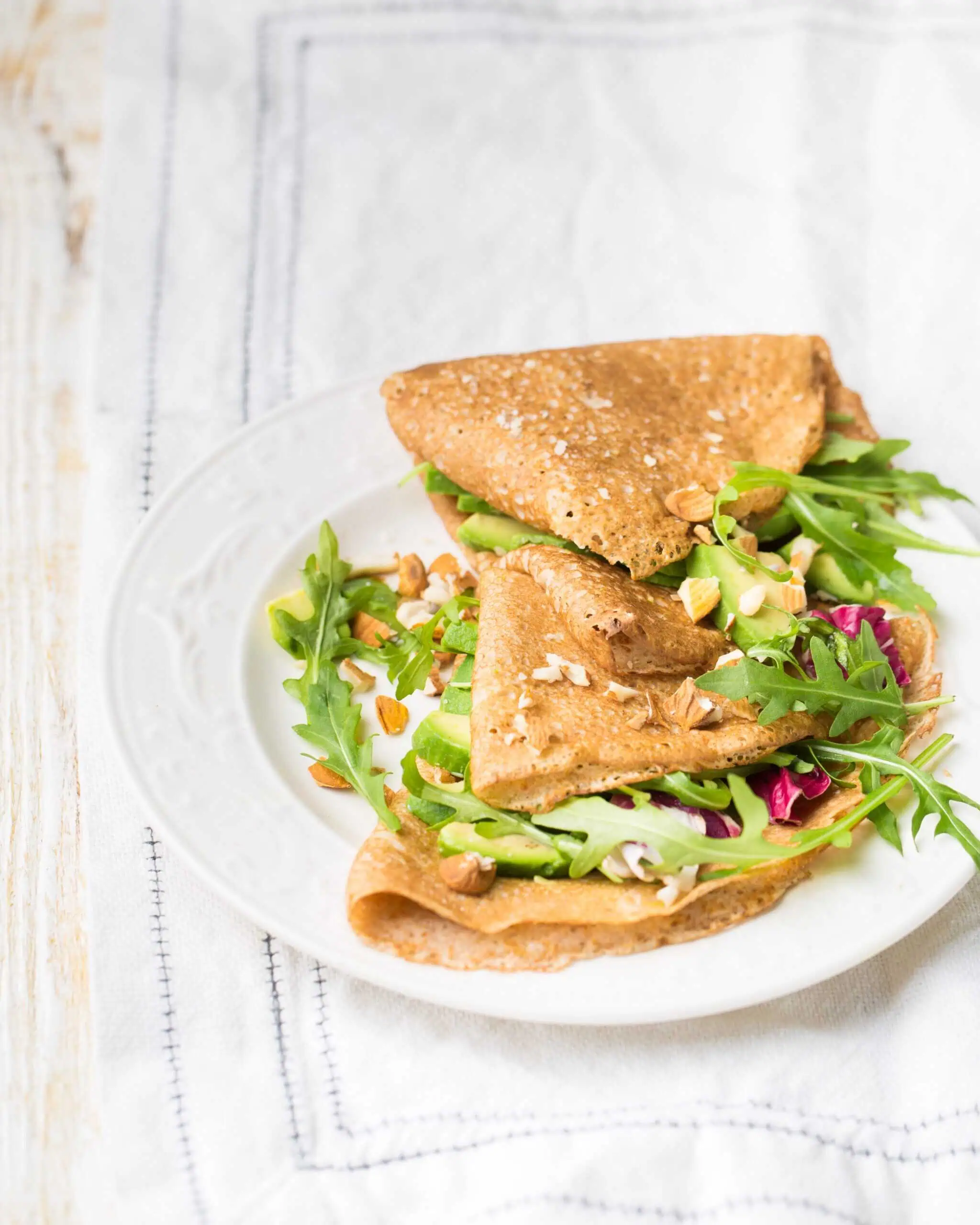 A white plate with two folded buckwheat crepes filled with arugula, mixed greens, and chopped nuts, placed on a white cloth.