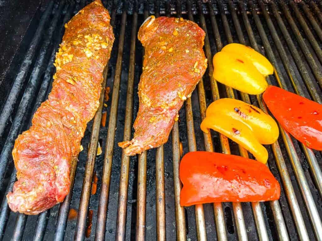 Hanger steaks on a grill with peppers nearby.