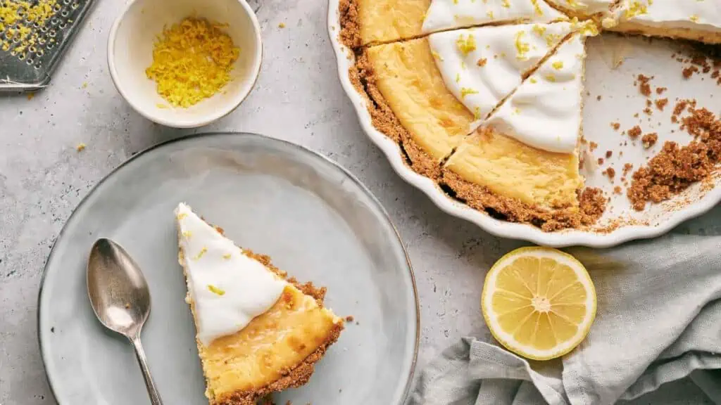 A slice of lemon pie on a plate with a spoon next to it and the remaining pie in a pie dish.