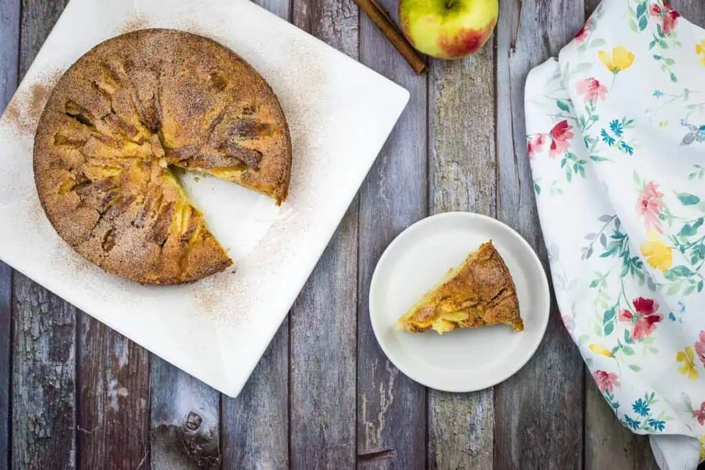 A slice of apple cake on a plate with cinnamon sticks.