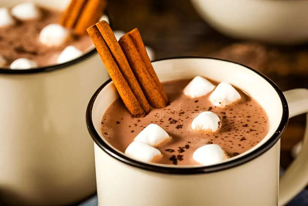 Two mugs of hot chocolate with cinnamon sticks and marshmallows.