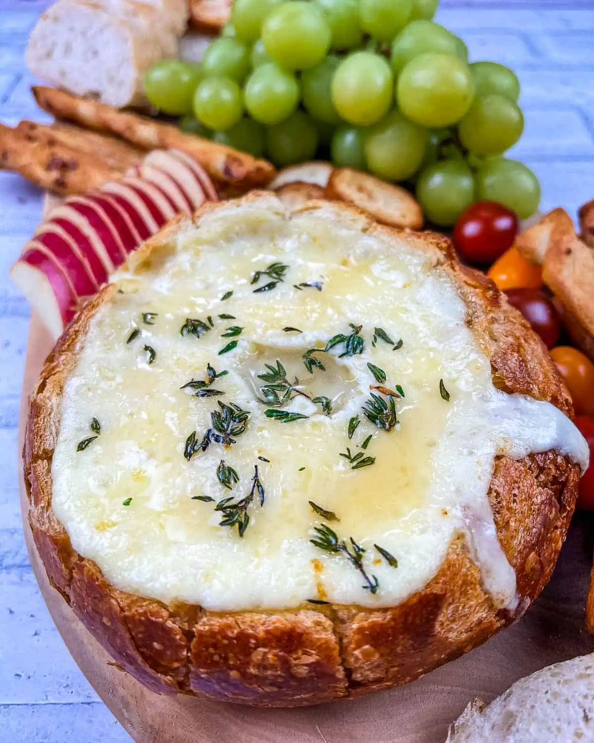 A wooden platter with baked brie in a bowl, cheese, crackers, grapes and bread.