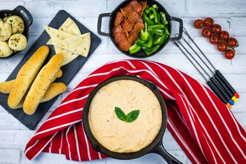 A bowl of pizza fondue with bread and breadsticks on a table.