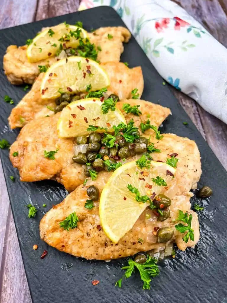 Chicken Picatta with lemon and parsley on a black plate.