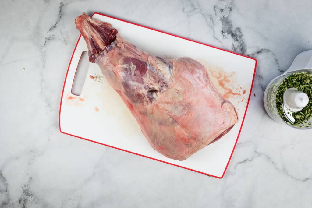 A leg of lamb on a cutting board next to a bowl of spices.