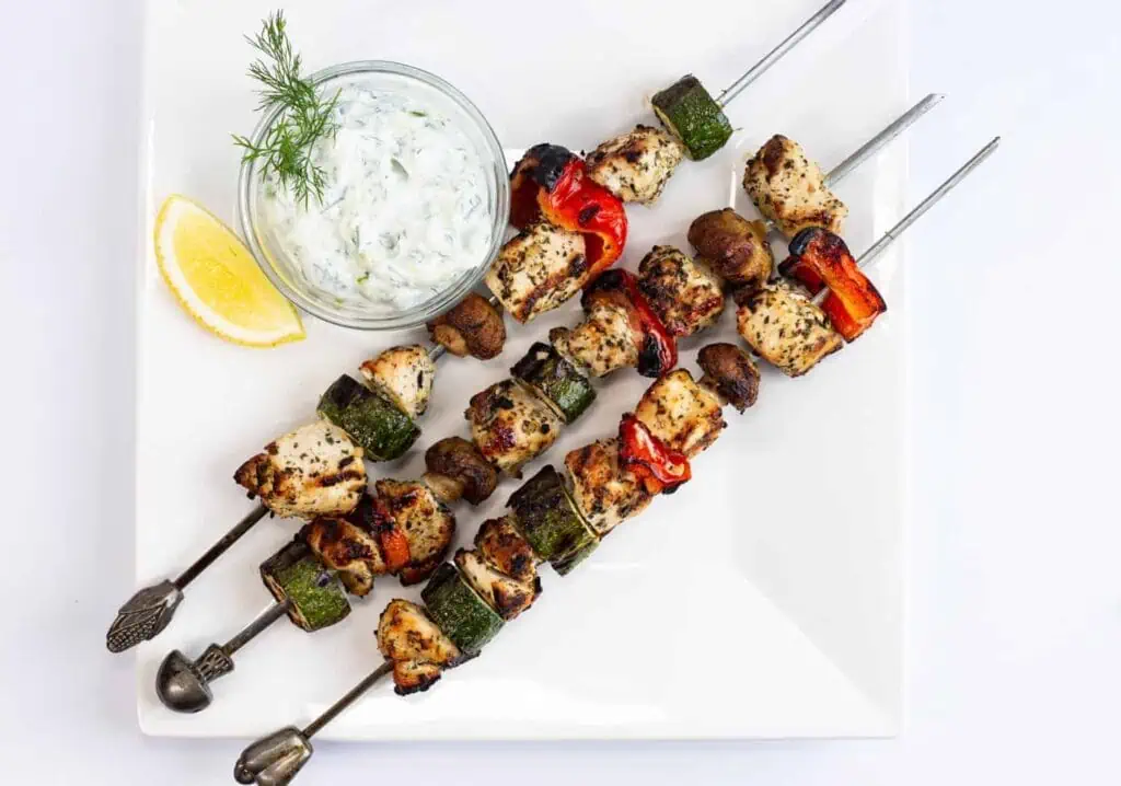 Grilled chicken skewers with tzatziki sauce on a white plate.