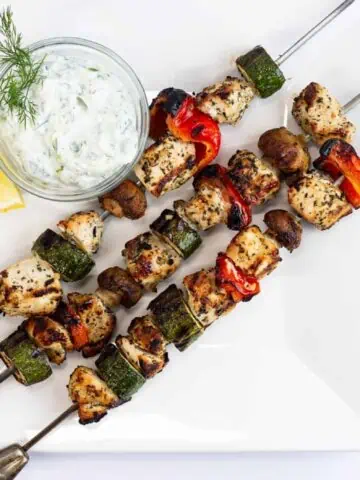 Grilled chicken skewers with tzatziki sauce on a white plate.