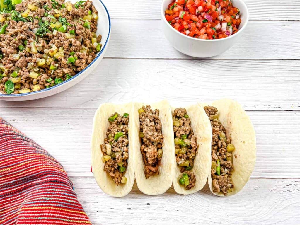 Four tacos with ground beef and vegetables on a white wooden surface, with additional fillings in bowls nearby.