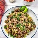 A plate of seasoned ground meat garnished with fresh herbs, accompanied by lime slices, garlic, and a bowl of diced vegetables.